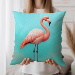 May include: A square decorative pillow featuring a flamingo. The flamingo is pink with a black beak and long legs, set against a turquoise background. The pillow is held by a person.