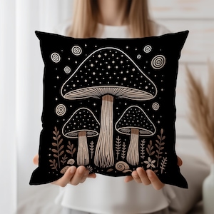 May include: Black square throw pillow featuring a whimsical mushroom design. The illustration includes three mushrooms with dotted caps and detailed stems, surrounded by botanical elements in shades of brown and beige. The pillow is held in front of a white background.