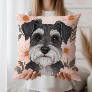 Könnte beinhalten: Ein dekoratives quadratisches Kissen mit einer detaillierten Illustration eines Schnauzer-Hundes mit schwarzem und grauem Fell, braunen Augen und einem weißen Bart. Das Kissen hat einen hellrosa Hintergrund mit pfirsichfarbenen Blumen und grünen Blättern.