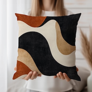 May include: Square decorative pillow with a modern abstract design. The pillow features a pattern of flowing shapes in black, beige, brown, and rust colors. The pillow is held by a person.