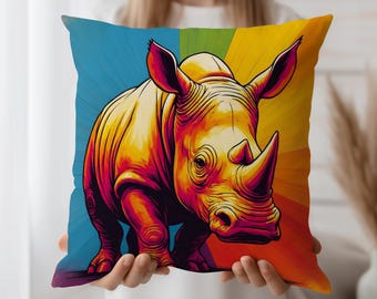 May include: Square throw pillow featuring a vibrant, stylized illustration of a rhinoceros. The rhino is rendered in warm tones of yellow, orange, and red, set against a rainbow-colored background. The pillow is held by a person.