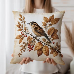 Puede incluir: Cojín decorativo cuadrado con una ilustración detallada de un pájaro posado en una rama con hojas de otoño y bayas rojas. El cojín tiene un aspecto vintage envejecido con un fondo crema y detalles en marrón y naranja.