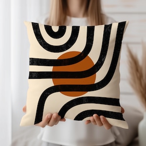 May include: Square decorative pillow with a cream background and a geometric design. The design features black curved lines, a burnt orange circle, and a textured appearance. The pillow is held by a person.