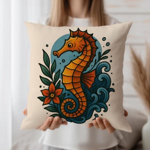 May include: A cushion with a colourful seahorse illustration. The seahorse is depicted in shades of orange and yellow, set against a blue backdrop with stylized waves and a flower. Green leaves and a red-orange flower add detail. The cushion is held by a person.
