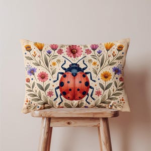 May include: Rectangular cushion with a colourful illustration of a ladybird with a red shell and black spots, set amongst flowers and foliage. The cushion has a beige background and is placed on a wooden stool. A charming accent piece for any room.