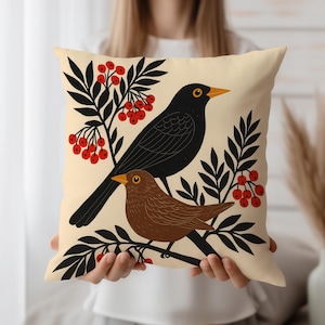 May include: Beige throw pillow featuring a black bird and a brown bird perched on a branch with red berries and black leaves. The birds have yellow beaks and eyes. The pillow is a square shape.