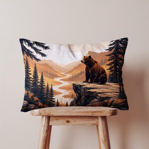 May include: Rectangular pillow featuring a brown bear sitting on a cliff overlooking a river winding through a mountain landscape. The artwork has a warm color palette of browns, oranges, and greens, with tall pine trees framing the scene.