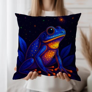Puede incluir: Cojín cuadrado con una vibrante ilustración de una rana azul con detalles en naranja y amarillo. La rana está sobre un fondo azul oscuro con estrellas y hojas. El cojín se muestra sobre un fondo blanco.