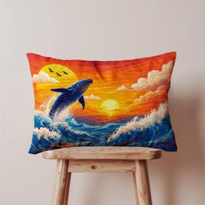 May include: Rectangular pillow featuring a vibrant sunset scene with a breaching blue whale. The artwork includes crashing waves, fluffy clouds, and silhouetted birds, set against a warm orange and yellow sky. The pillow is placed on a wooden stool.