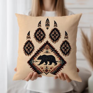 Op de afbeelding: Een beige vierkant kussen met een zwart berenpootafdrukontwerp. De pootafdruk bestaat uit geometrische patronen in bruin, zwart en blauwgroen. Een zwart berensilhouet is gecentreerd in een ruitvormig patroon.