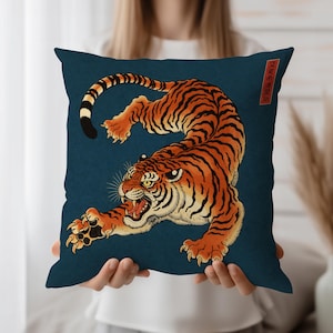以下が含まれることがあります： 濃い青色の背景に、オレンジと黒の虎が描かれた正方形のクッション。 虎はダイナミックなポーズで、口を開き、爪を伸ばしています。 右上には日本語のテキストがあります。