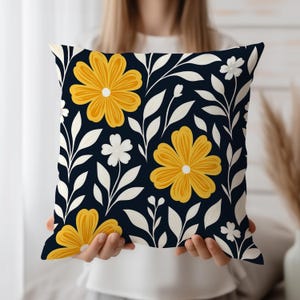 Op de afbeelding: Vierkant decoratief kussen met een bloemenpatroon. Het heeft grote gele bloemen met witte centra, witte bladeren en kleine witte bloemen tegen een donker marineblauwe achtergrond. Het kussen wordt door een persoon vastgehouden.