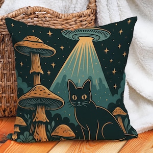 May include: Cushion with a fantasy design featuring a black cat, mushrooms, and a UFO. The pillow has a dark teal background with gold and tan accents. The cat is sitting in front of the mushrooms.