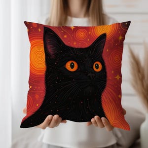 May include: Square decorative pillow featuring a black cat with bright orange eyes. The cat is set against a vibrant red background with yellow spiral and star patterns. The pillow is held in front of a person.