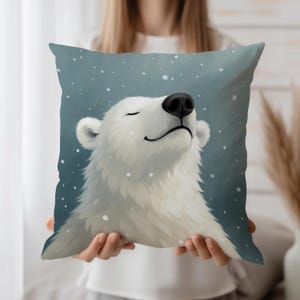 Könnte beinhalten: Quadratisches Zierkissen mit einem lächelnden Eisbären mit geschlossenen Augen, vor einem blauen Hintergrund mit fallenden Schneeflocken. Der Bär hat weißes Fell und eine schwarze Nase. Ein dekorativer Artikel für ein Wohn- oder Schlafzimmer.