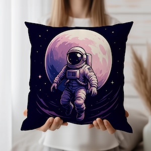 Op de afbeelding: Een decoratief sierkussen met een astronaut die in de ruimte zweeft. De astronaut draagt een ruimtepak met een Amerikaanse vlag patch, tegen een achtergrond van een grote, roze en paarse maan en een donkerblauwe sterrenhemel.