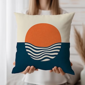 May include: A decorative pillow with a minimalist design. The top half features a cream-colored background with a large orange sun. The bottom half is a deep blue with white wavy lines, representing the ocean. A simple, modern home decor item.