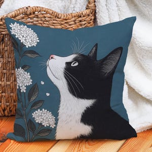 Op de afbeelding: Vierkant kussen met een tealblauwe achtergrond met een zwart-witte kat die naar witte bloemen kijkt. De kat heeft een roze neus en staat tegen een bloemenontwerp. Het kussen ligt op een houten oppervlak.