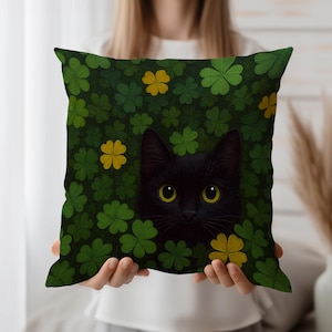 Könnte beinhalten: Ein quadratisches Zierkissen mit einer schwarzen Katze, die aus einem Feld aus grünen und gelben Kleeblättern hervorschaut. Die Katze hat gelbe Augen und ein schwarzes Fell. Das Kissen wird von einer Person gehalten.