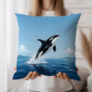 Puede incluir: Un cojín cuadrado con una ilustración de una orca saltando. El diseño muestra una orca en blanco y negro rompiendo el agua, con un cielo azul y un fondo oceánico. El cojín es sostenido por una persona.