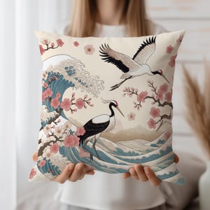 Puede incluir: Cojín cuadrado decorativo con un diseño de inspiración japonesa. Muestra dos grullas, flores de cerezo y olas estilizadas en tonos azules, rosas y beige. La obra de arte evoca serenidad y belleza natural.
