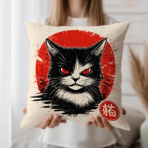 Op de afbeelding: Een decoratief kussen met een zwart-witte kat met rode ogen tegen een rood zonontwerp. Het kussen heeft een crèmekleurige achtergrond en een Japans karakter in een rode cirkel.