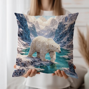Puede incluir: Cojín cuadrado decorativo con un oso polar en un paisaje montañoso nevado. El oso es blanco, el agua turquesa y las montañas azules y blancas. El cojín está sostenido por una persona.