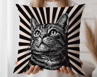 May include: A decorative square pillow featuring a detailed black and white illustration of a cat's face. The cat is looking upwards, set against a backdrop of black and cream-colored radial stripes. The pillow is held by a person.