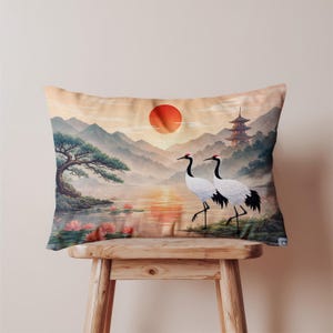 以下が含まれることがあります： 赤い太陽、山、塔のある穏やかな日本の風景が描かれた長方形のクッション。 黒い翼の先端と赤い王冠を持つ2羽の白い鶴が湖の近くに立っています。 クッションは木製のスツールの上に置かれています。