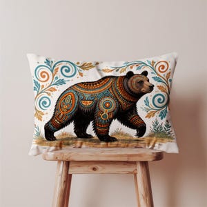 May include: Decorative rectangular pillow featuring a walking bear with intricate patterns in orange, teal, and brown. The bear is set against a white background with floral accents. The pillow is placed on a wooden stool.