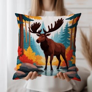 Puede incluir: Un cojín cuadrado decorativo con una escena de bosque que representa un alce marrón. La obra de arte incluye árboles coloridos, rocas y agua, con una paleta de marrón, azul, amarillo y rojo. El cojín es sostenido por una persona.
