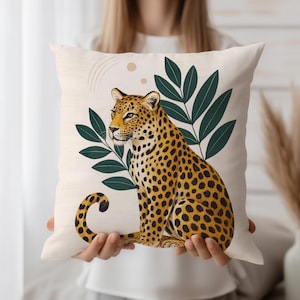 May include: Square decorative pillow with a leopard illustration. The pillow features a sitting leopard with yellow and black spots, green leaves, and beige background. The pillow is held by a person.