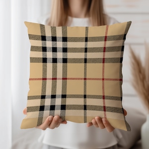 Può includere: Un cuscino quadrato con sfondo beige e motivo a quadri neri, rossi e bianchi. Il cuscino è tenuto da una persona. Il design a quadri presenta linee intersecanti, creando un look classico. Un accessorio decorativo per la casa.