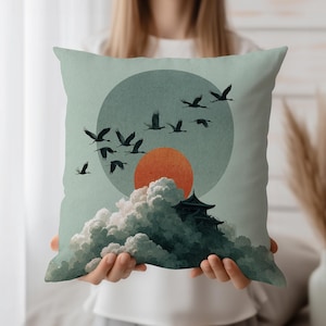 May include: Square decorative pillow featuring a Japanese-inspired design. The artwork depicts a flock of birds flying in front of a large orange sun, with a temple atop a mountain of clouds. The background is a muted teal color.