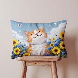May include: Rectangular pillow featuring a smiling orange and white cat in a field of sunflowers and blue flowers. White butterflies float in the sky. The pillow is resting on a wooden stool.