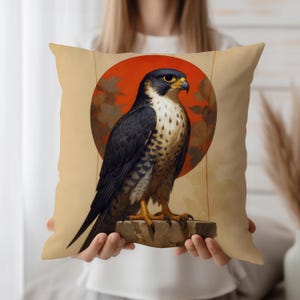 May include: A decorative pillow featuring a detailed illustration of a falcon perched on a stone. The bird has dark blue and white feathers, a yellow beak, and is set against a red circle and beige background. The pillow is square.