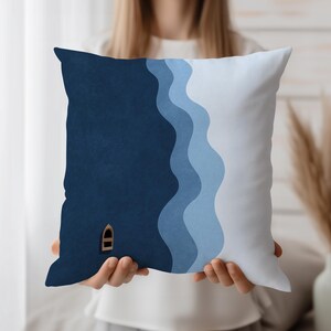 Puede incluir: Cojín cuadrado con diseño náutico. El cojín presenta un fondo azul oscuro con un pequeño barco de madera y rayas onduladas azules y celeste. El cojín está sostenido por una persona.