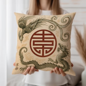 May include: Beige square pillow featuring a phoenix and dragon design with a central red emblem. The artwork is in a traditional Asian style, with detailed line drawings and a vintage aesthetic. The pillow is held by a person.