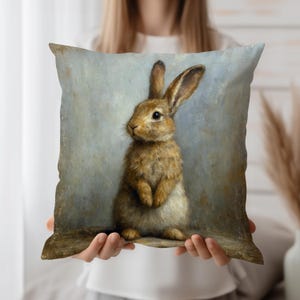May include: Square decorative pillow featuring a detailed painting of a brown rabbit with long ears. The rabbit is standing upright against a muted blue and gray background. The pillow has a vintage, artistic aesthetic.