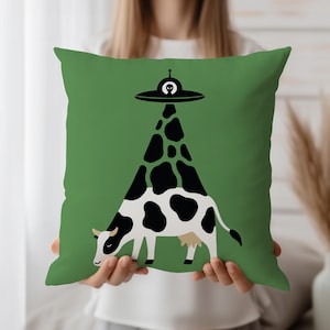May include: Green square cushion with a whimsical design featuring a black and white cow being abducted by a UFO. The UFO is black with a small alien figure. The cow's spots form a triangular shape.