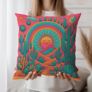 Könnte beinhalten: Ein quadratisches Zierkissen mit einem lebendigen Wüstenlandschaftsdesign. Das Kissen zeigt eine lächelnde Sonne, einen Regenbogen, Kakteen und Berge in den Farbtönen Pink, Orange, Türkis und Gelb. Das Kissen wird von einer Person gehalten.
