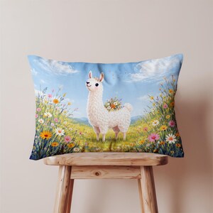 Puede incluir: Cojín rectangular con una ilustración caprichosa de una llama blanca adornada con flores, de pie en un campo de flores silvestres de colores. El fondo muestra un cielo azul con nubes. El cojín está sobre un taburete de madera.