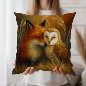 Op de afbeelding: Vierkantig decoratief kussen met een warmgekleurde illustratie van een vos en een uil met gesloten ogen, die tegen elkaar aan liggen. De vos heeft oranje vacht en de uil heeft bruine en witte veren. De achtergrond heeft een gouden tint.