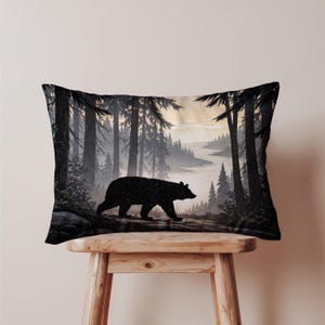 May include: Rectangular pillow featuring a black silhouette of a bear walking through a forest. The design includes tall trees, a river, and a muted color palette of black, gray, and beige. The pillow is placed on a wooden stool.