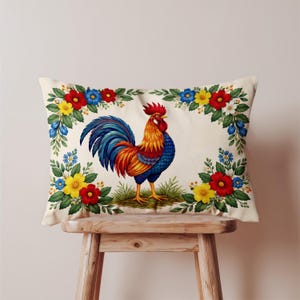 Può includere: Cuscino rettangolare decorativo con un'illustrazione colorata di un gallo. Il gallo è raffigurato con piume blu, arancioni e rosse vivaci, in piedi sull'erba verde. Il cuscino è incorniciato da un bordo floreale con fiori rossi, gialli e blu.