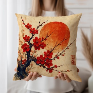 May include: Square cushion with a Japanese-style design. It features a red sun and a blossoming tree with red flowers on a beige backdrop. The design includes Japanese text in the lower right corner. Measurements are approximately 40cm x 40cm.