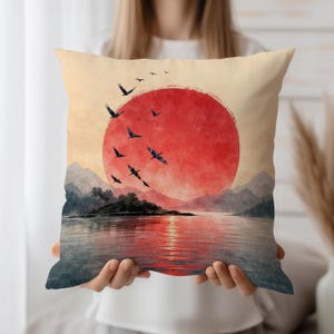 Op de afbeelding: Een decoratief kussen met een serene landschap. Een grote rode zon gaat onder boven het water, met vogels in vlucht. Het ontwerp is in aquarel stijl, met een beige achtergrond en een weerspiegeling van de zon in het water.
