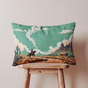 May include: Rectangular throw pillow with a vintage Western scene. The pillow features a cowboy on horseback, a desert landscape with cacti, and a small cabin. The color palette includes teal, brown, and orange tones.