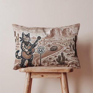 May include: A decorative pillow featuring a sepia-toned illustration of a cat playing a guitar on Route 66. The cat wears a bandana and cowboy boots, with a desert landscape and a vintage car in the background.