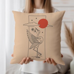 May include: Beige square cushion featuring a black line drawing of a crane holding a flower, with a red sun and clouds. The crane is inside a rectangular frame. The cushion is held by a person.